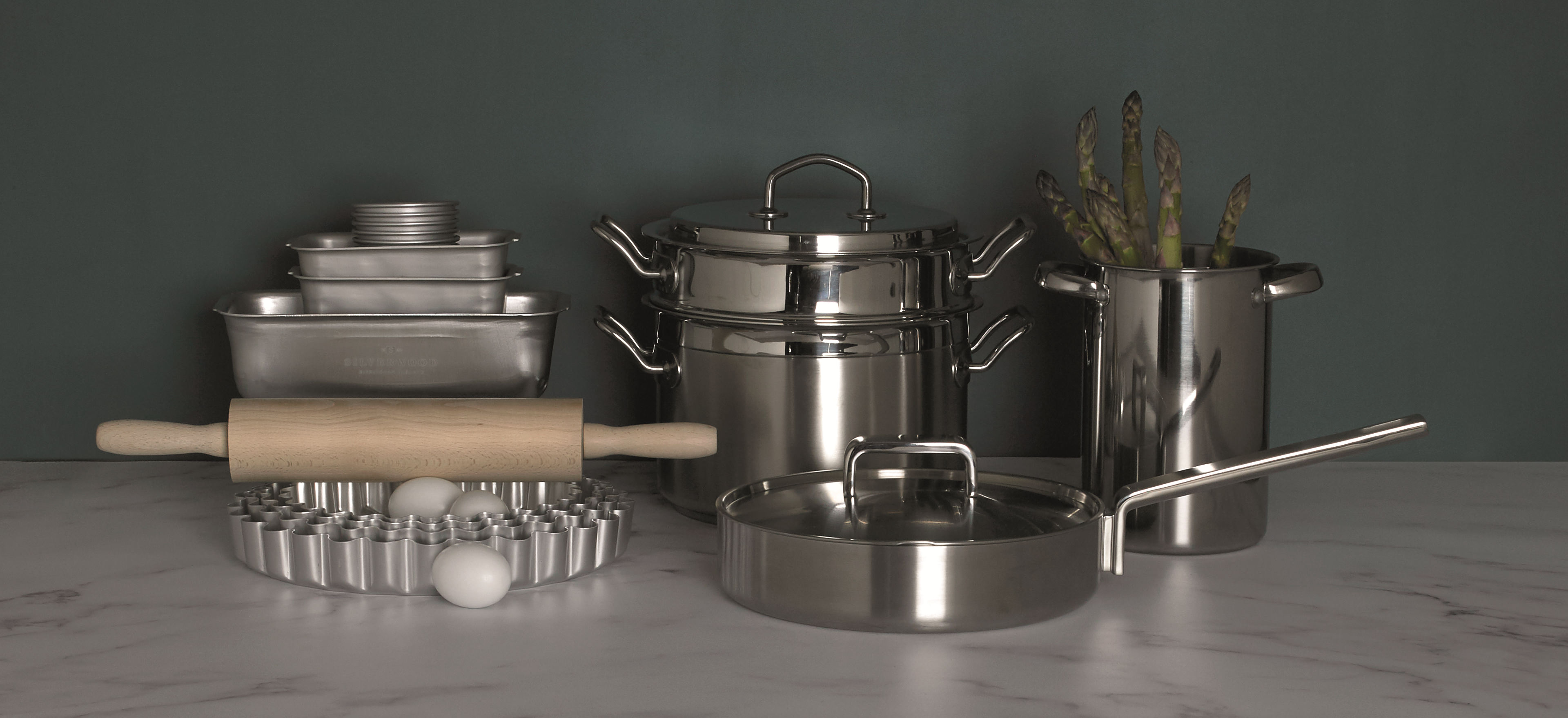 Pots and pans from our cookware ranges and bakeware items such as a rolling pin and flan tin arranged on a turquoise background.