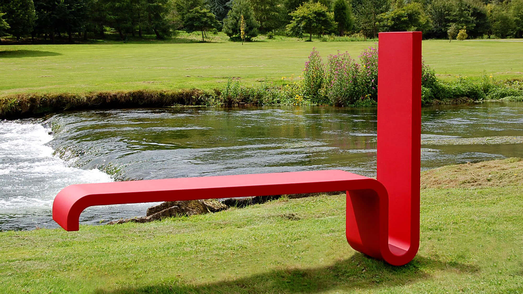 Red bench in situ on grass next to a stream.