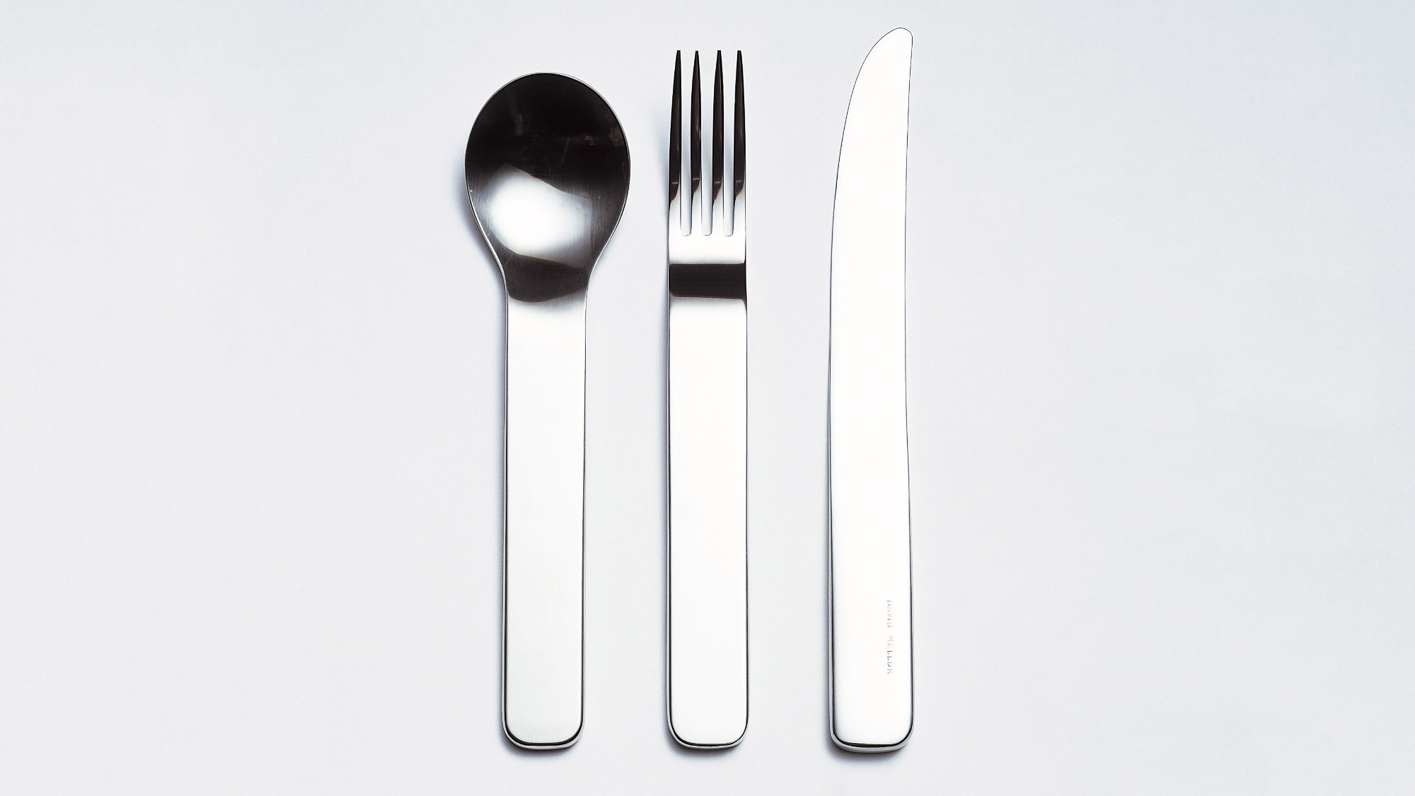 Minimal stainless steel cutlery items displayed in a line on a grey background.
